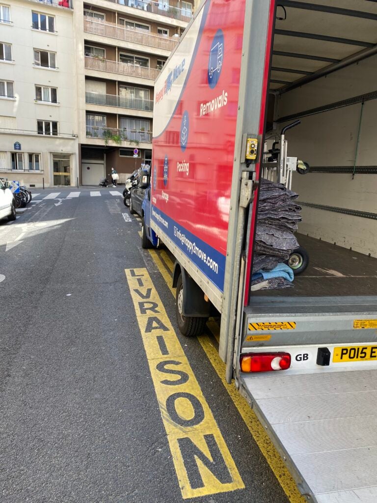 A delivery truck with branding for a moving company is parked on a city street in Paris, marked with "LIVRAISON." The truck is partially loaded with moving blankets and a dolly, ready to assist in relocating to a cozy 1-bedroom flat. Apartment buildings rise majestically in the background.
