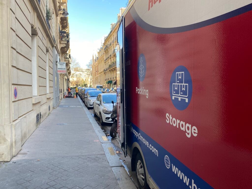 A moving truck is parked on a narrow Parisian street alongside cozy sidewalks. The truck showcases graphics for packing and storage services. Cars are lined neatly along the road, with historic buildings flanking both sides under a clear blue sky.