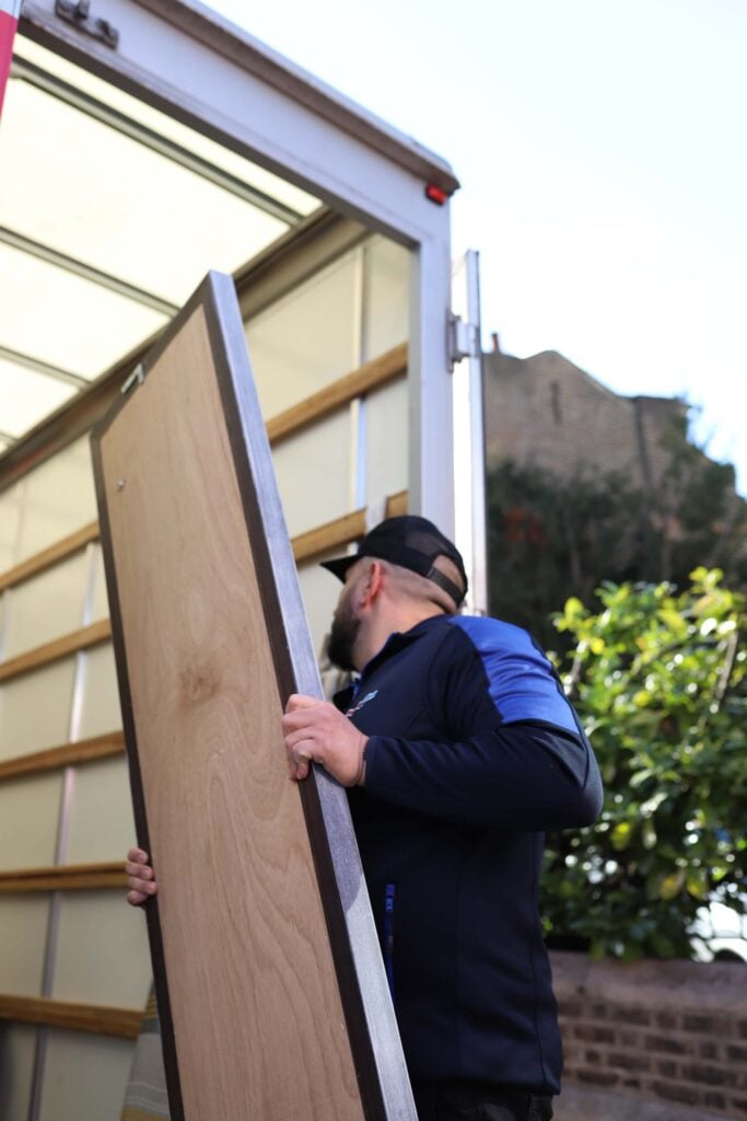 A man in a black cap and blue jacket is packing a wooden door onto a moving truck, perhaps destined for a new 6-bedroom house. The truck's rear is open, revealing glimpses of a green plant and building in the sunlit background.