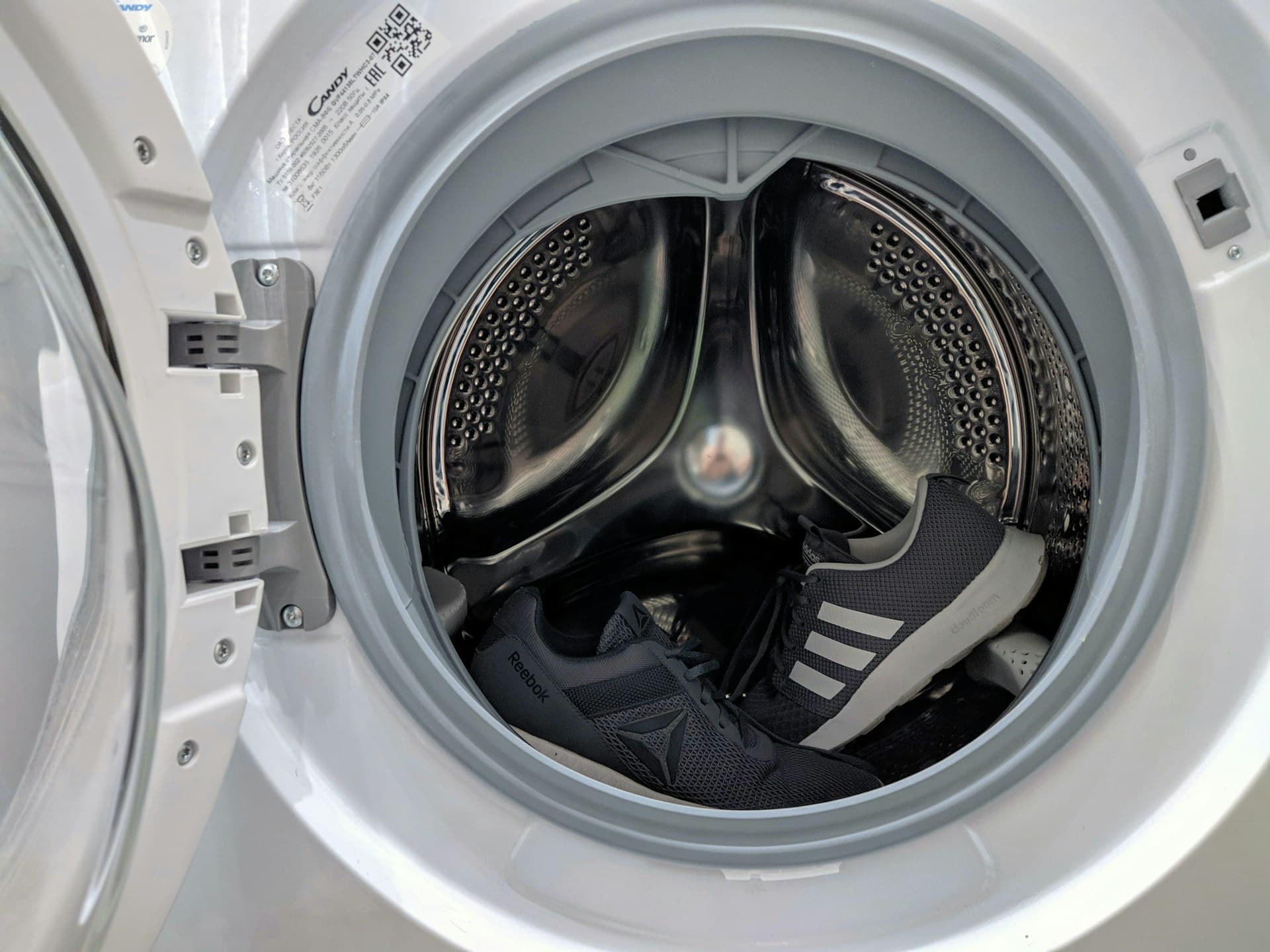 The washing machine's door is open, ready for action, with two black and white athletic shoes nestled inside the drum. As you prepare the washing machine, the shoes sit side by side, surrounded by the pristine interior waiting to unleash its cleaning magic.