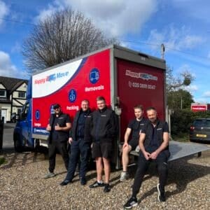 Five men pose in front of a Happy2Move removals truck parked on a gravel driveway, with houses and trees in the background on a sunny day, showcasing the team's dedication to their jobs.