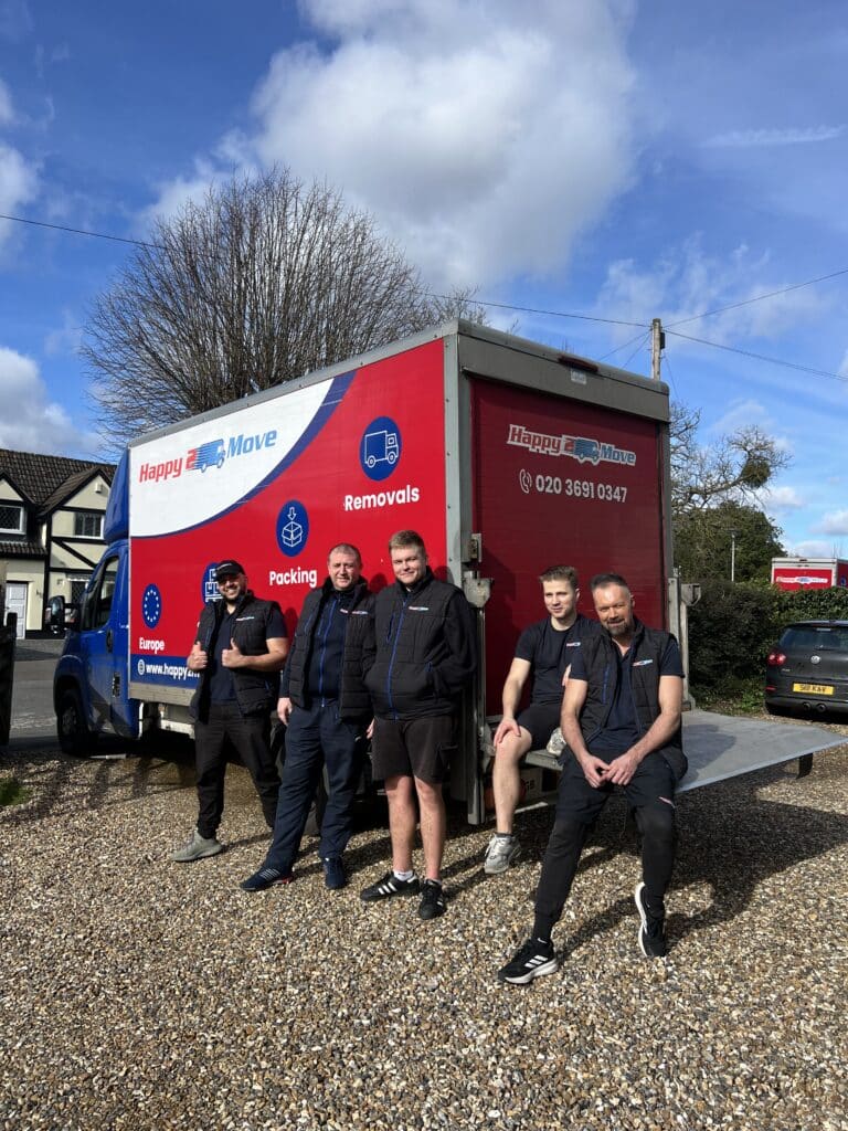 Five men pose in front of a Happy2Move removals truck parked on a gravel driveway, with houses and trees in the background on a sunny day, showcasing the team's dedication to their jobs.