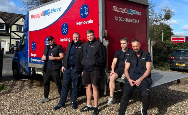 Five men pose in front of a Happy2Move removals truck parked on a gravel driveway, with houses and trees in the background on a sunny day, showcasing the team's dedication to their jobs.