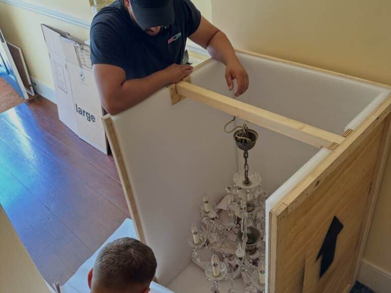 Two people carefully pack a glass chandelier inside a custom wooden crate lined with foam for protection, showcasing the care and precision required in flatpack assembly. A large cardboard box sits on the hardwood floor in the background.