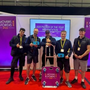 Five men stand smiling on a red carpet at an awards event, holding trophies and a branded moving dolly. The backdrop displays “Movers & Storers Show 2025” and “Packer of the Year Presentation.”.