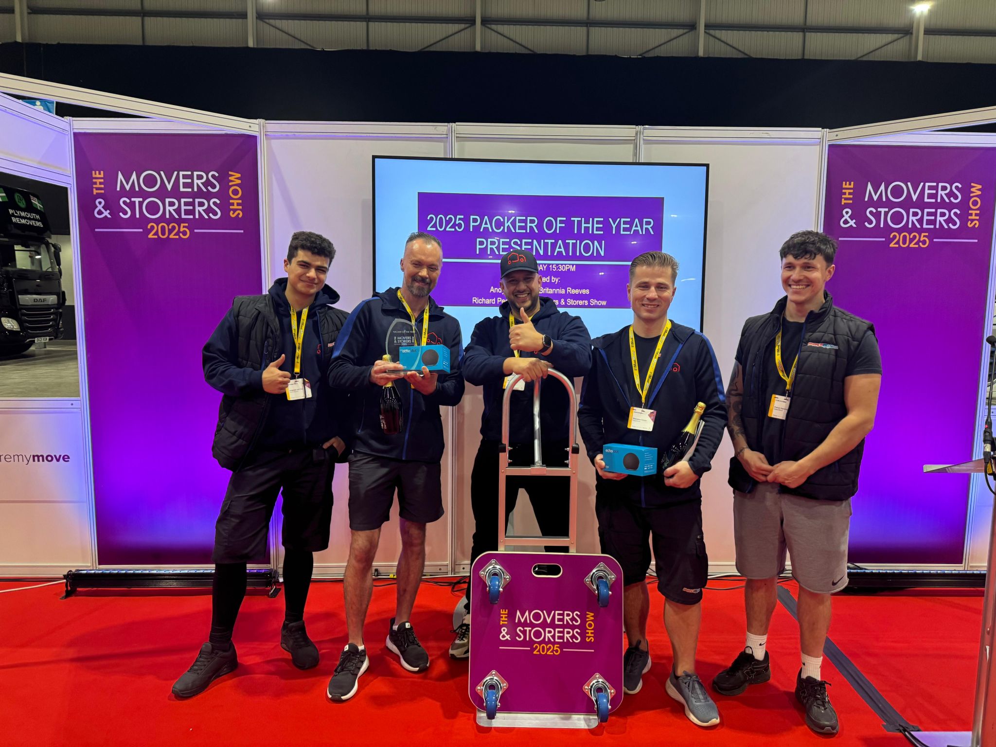 Five men stand smiling on a red carpet at an awards event, holding trophies and a branded moving dolly. The backdrop displays “Movers & Storers Show 2025” and “Packer of the Year Presentation.”.