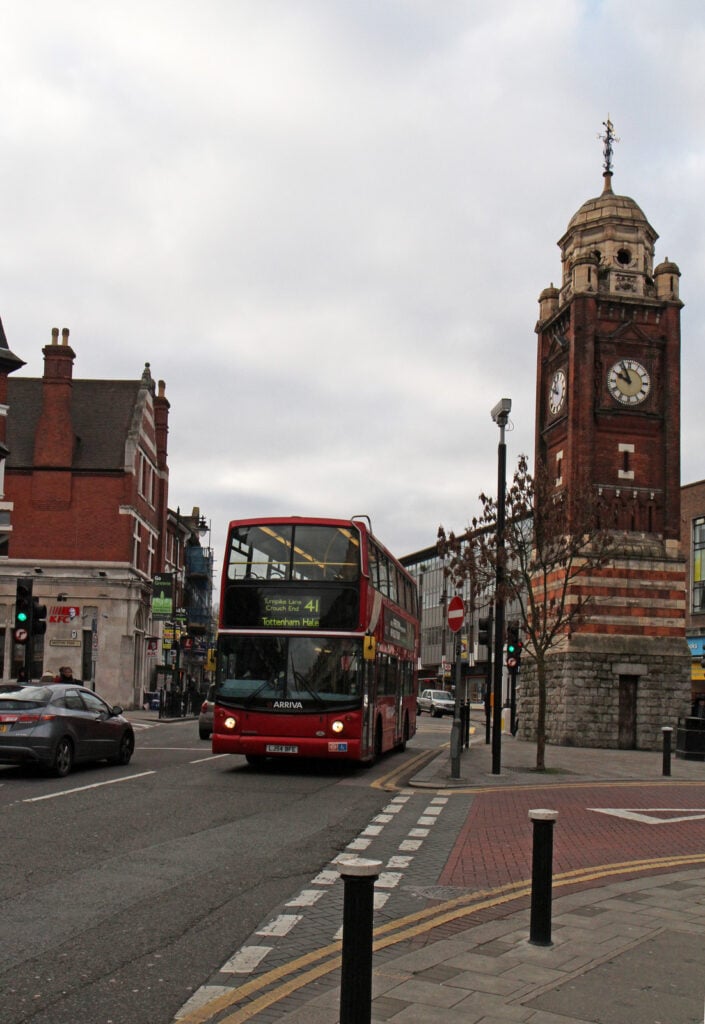 A red double-decker bus drives past a historic clock tower on a city street lined with brick buildings, hinting at popular areas in London to explore for those moving to London, all under a cloudy sky.