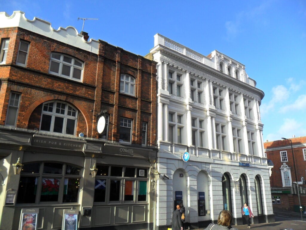 Street view of two contrasting buildings in one of the best places London has to offer: a red-brick Irish pub with flags on the left and a grand white classical building on the right, under a clear sky with a few people walking by.