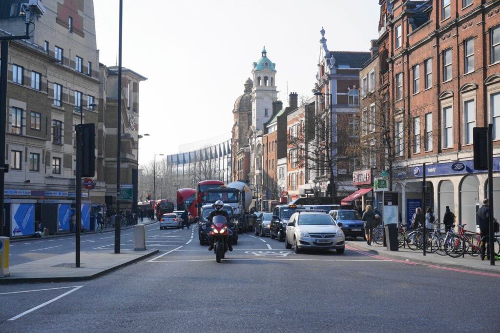 A busy city street in one of the popular areas London offers, with cars, buses, and a motorbike in motion. People walk along the pavements, and bicycles are parked beside red-brick buildings under a clear sky.