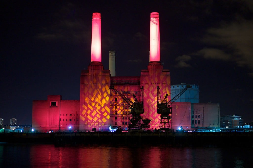 A large industrial building with four tall chimneys, located in one of the popular areas of London, is illuminated at night with vibrant pink and orange lights, reflecting on the water beside cranes under a dark, cloudy sky.