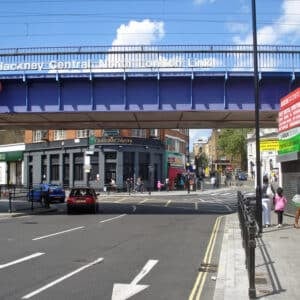 Street view of Hackney Central, one of the popular areas London offers, with a blue railway bridge overhead, cars on the road, pedestrians strolling, and shops like the Railway Tavern on the corner. Bright, sunny weather.