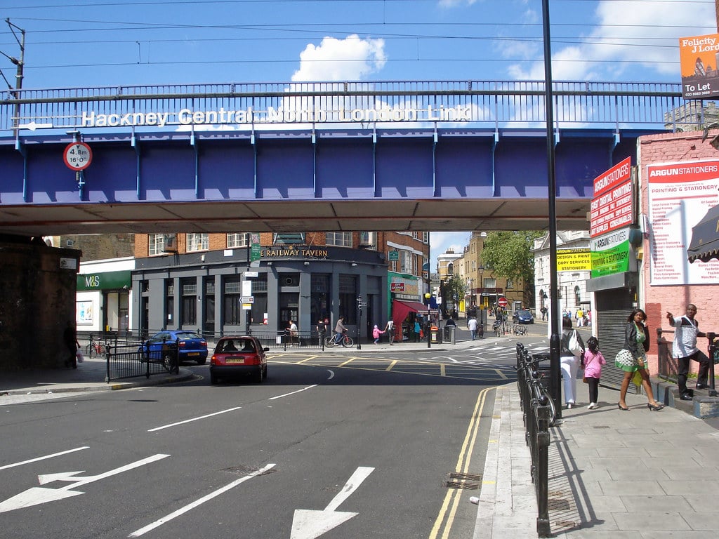 Street view of Hackney Central, one of the popular areas London offers, with a blue railway bridge overhead, cars on the road, pedestrians strolling, and shops like the Railway Tavern on the corner. Bright, sunny weather.