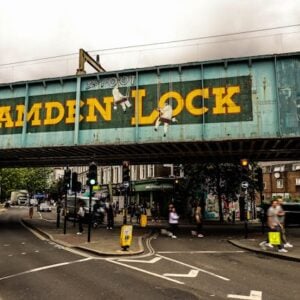 A painted railway bridge with large yellow letters reading "Camden Lock" spans a busy street in one of the hardest to move London boroughs, with pedestrians, cars, and shops visible beneath a cloudy sky.