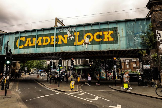 A painted railway bridge with large yellow letters reading "Camden Lock" spans a busy street in one of the hardest to move London boroughs, with pedestrians, cars, and shops visible beneath a cloudy sky.