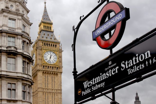 A view of the Westminster Station Underground sign in London, with the iconic Big Ben clock tower and part of a historic building visible in the background under a cloudy sky—one of the most recognisable sights across all London boroughs.