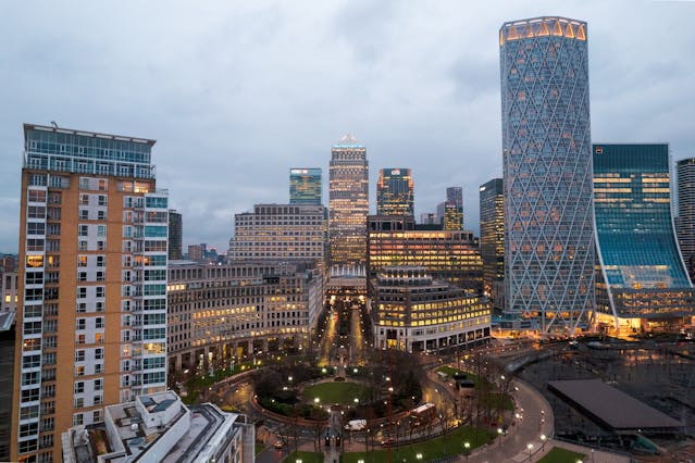 A cityscape at dusk shows modern high-rise buildings with lights on, surrounding a small park with pathways and trees. The cloudy sky frames both residential and office towers, typical of London boroughs known for their moving difficulty.