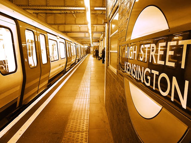 A London Underground train at High Street Kensington station, one of the London boroughs often noted for difficulty in moving, with the station sign visible on the right and passengers waiting on the platform under yellow-tinted lighting.
