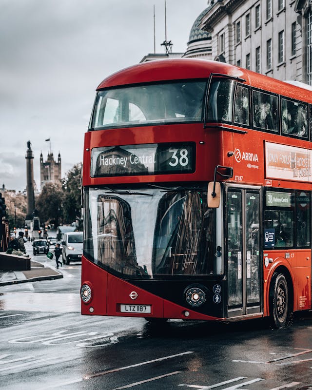 A red double-decker bus on a wet London street displays route 38 to Hackney Central, one of the London boroughs often considered among the hardest to move through. Buildings and pedestrians are visible in the background under a cloudy sky.