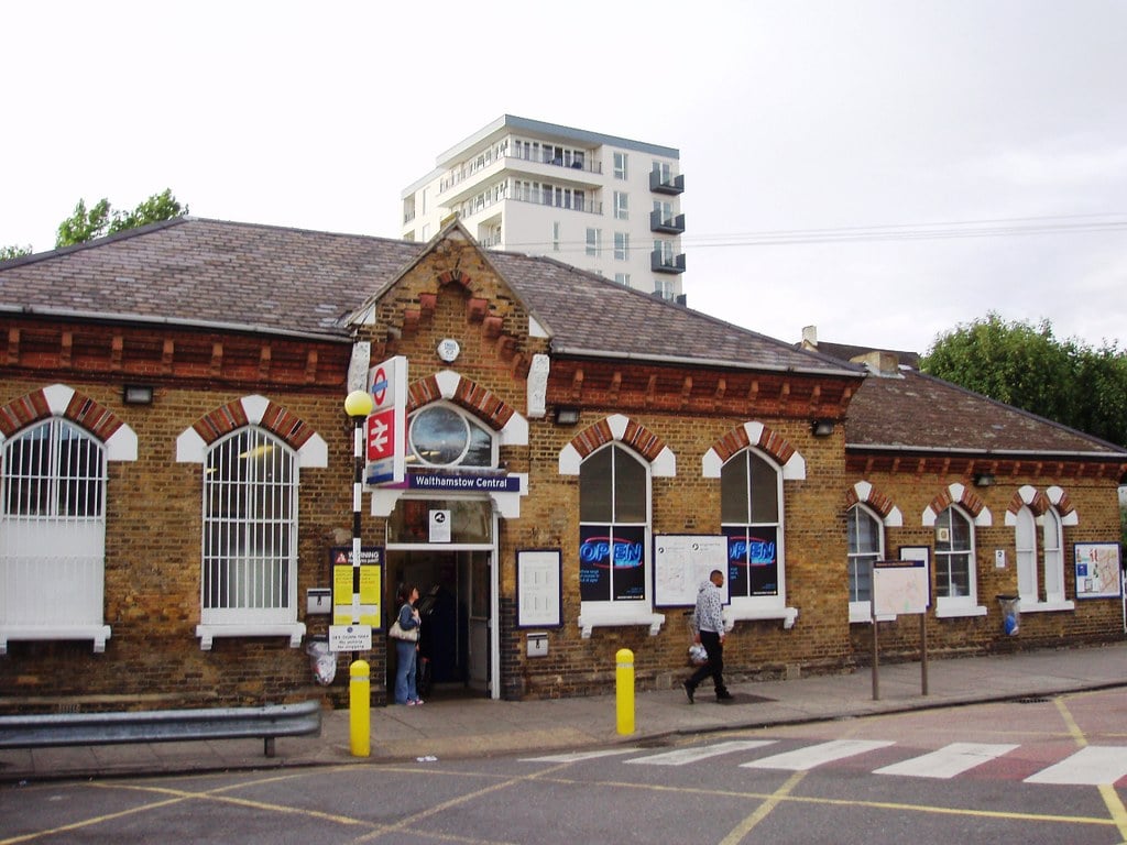 A brick railway station building with arched windows and a sign reading "Walthamstow Central." People walk near the entrance; a modern block of flats rises in the background—a glimpse of one of the popular areas in London.