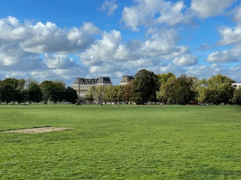 A wide, grassy park under a partly cloudy blue sky, with trees and several large buildings in the background. A few people are visible in the distance enjoying the open space—a perfect spot for anyone considering moving to Clapham.