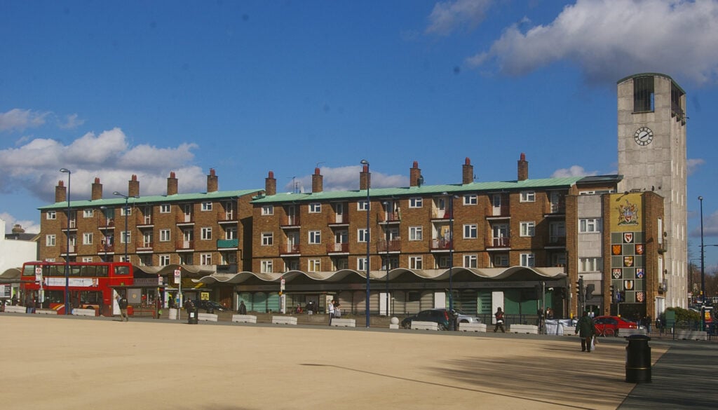 A wide view of a square in Walthamstow, with brick buildings, a clock tower, a red double-decker bus, and people and cars under a blue sky—a charming scene from any moving to Walthamstow guide.