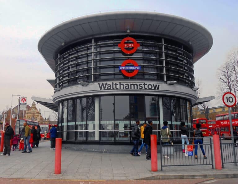 The modern circular glass entrance of Walthamstow station, featured in this Walthamstow guide, displays "Buses" and "Underground" roundels above its name. People walk outside, with red double-decker buses in the background.