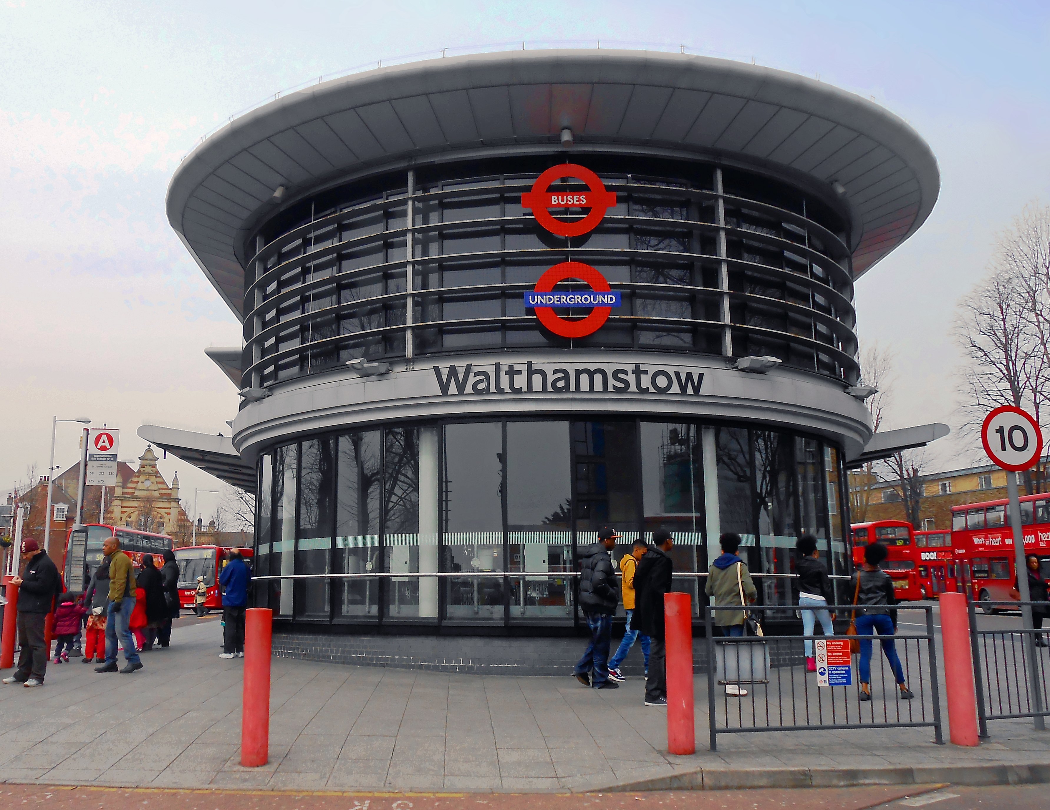 The modern circular glass entrance of Walthamstow station, featured in this Walthamstow guide, displays "Buses" and "Underground" roundels above its name. People walk outside, with red double-decker buses in the background.