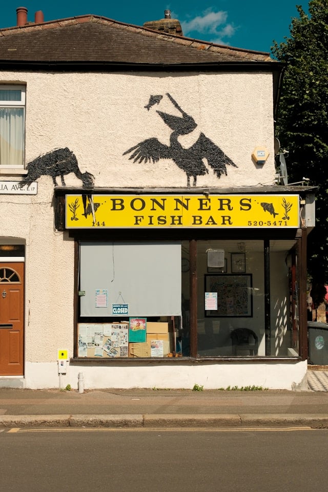 The exterior of Bonners Fish Bar in Walthamstow features a bright yellow sign, black bird silhouettes above it, and a large closed window facing the street—a classic spot to guide you when moving through the area.