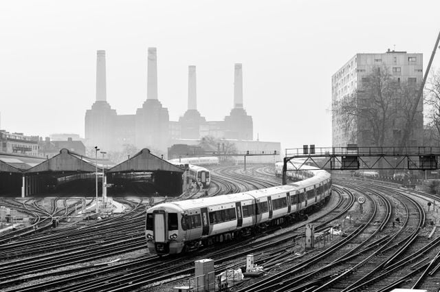 A modern train glides along curved railway tracks in an urban setting, with multiple tracks converging. In the misty background, Battersea’s iconic industrial building with four tall chimneys rises—perfect for a Battersea guide or those moving to Battersea.