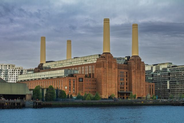 A large brick building with four tall white chimneys stands by the water, surrounded by modern glass buildings under a cloudy sky—a striking sight for anyone considering moving to Battersea or seeking a local Battersea guide.