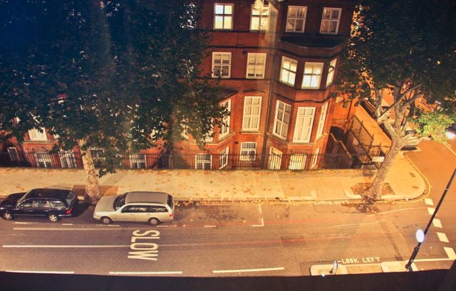Night-time view from above of a street in Clapham with parked cars, a pavement lined with trees, and a brick building. The road has "SLOW" and "LOOK LEFT" painted on it—perfect for anyone considering moving to Clapham.