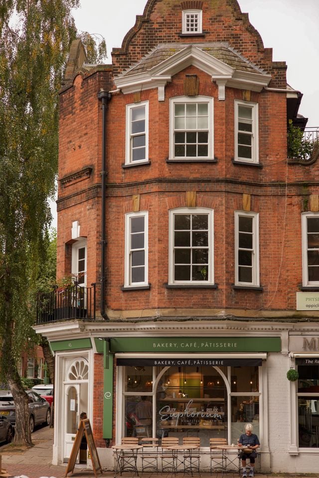 A red-brick building with white-trimmed windows houses Superbon, a bakery, café, and patisserie—a local favourite for those moving to Clapham. Outdoor tables and chairs invite you to relax among trees and parked cars nearby.