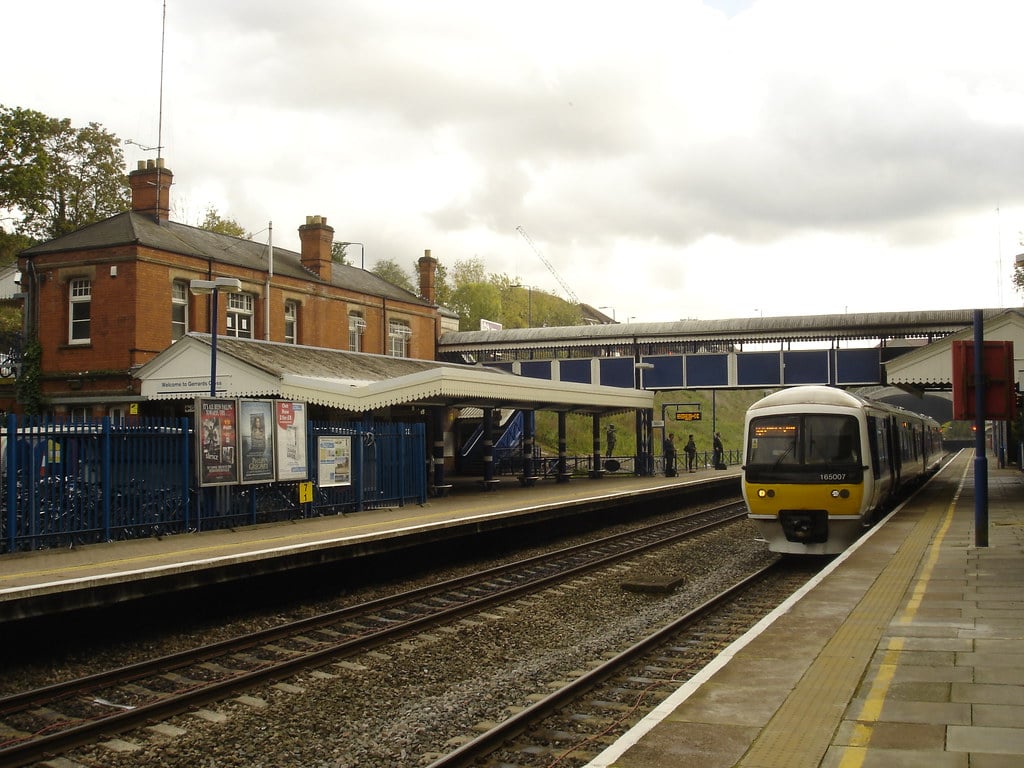 A moving train arrives at a small, cloudy railway station with a red brick building, blue fences, and a footbridge connecting the platforms—much like the charming atmosphere of Gerrards Cross during removal.