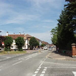 A quiet suburban street in Gerrards Cross with trees, buildings, and parked cars on both sides. The sky is clear and blue, and a few people are visible walking along the pavements—perhaps discussing a recent relocation or planning their next move.
