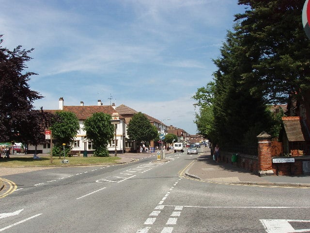 A quiet suburban street in Gerrards Cross with trees, buildings, and parked cars on both sides. The sky is clear and blue, and a few people are visible walking along the pavements—perhaps discussing a recent relocation or planning their next move.