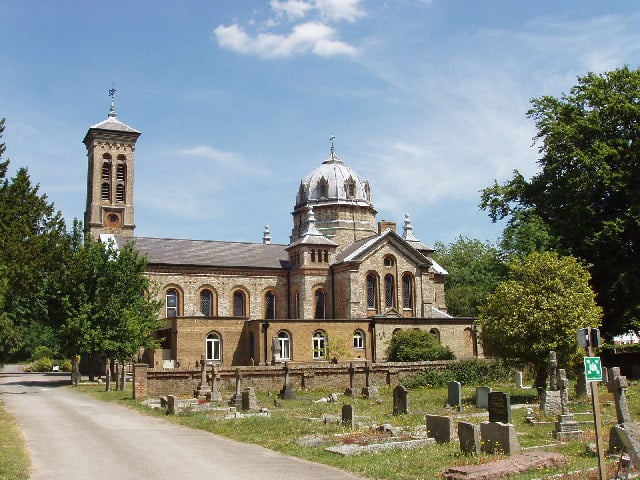 A historic church with a central dome and tall bell tower stands beside a graveyard on a sunny day in Gerrards Cross, with gravestones and greenery in the foreground.