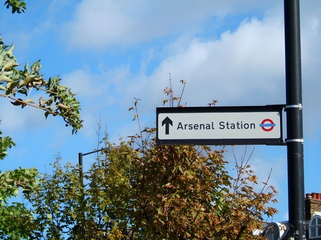 A sign with an arrow and the London Underground logo points towards Arsenal Station, surrounded by trees under a partly cloudy blue sky—a familiar sight for anyone moving to Islington or considering an Islington relocation.