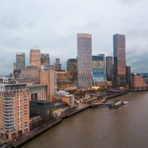 A cityscape of Canary Wharf in London, featuring modern skyscrapers and buildings along the River Thames under a cloudy sky at dusk—a striking view that captures the vibrant atmosphere for anyone considering moving to Canary Wharf.