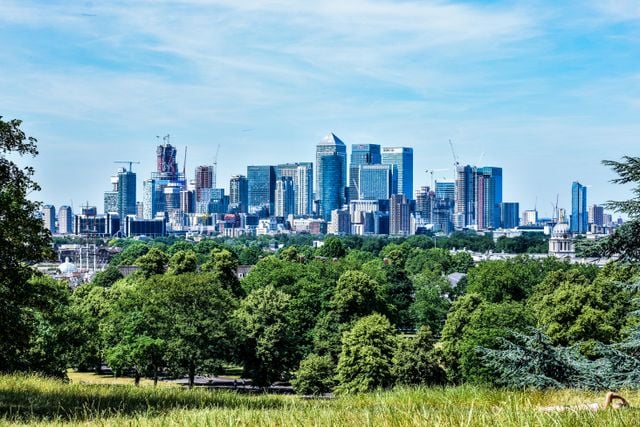 A city skyline with modern skyscrapers rises above a lush, green park filled with trees under a blue sky. Construction cranes are visible among the buildings, reflecting the dynamic growth and vibrant atmosphere that draws many to consider living in Canary Wharf.