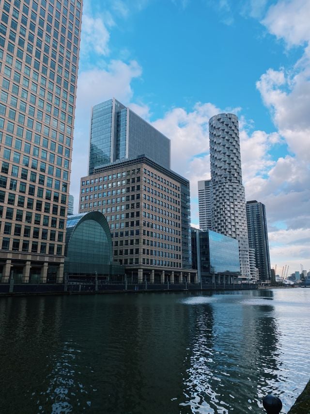 Modern high-rise buildings, including some with unique geometric designs, are reflected in a calm river under a blue, partly cloudy sky. In the foreground, water fountains add to the scenic beauty—an inviting view for anyone considering relocation to Canary Wharf.