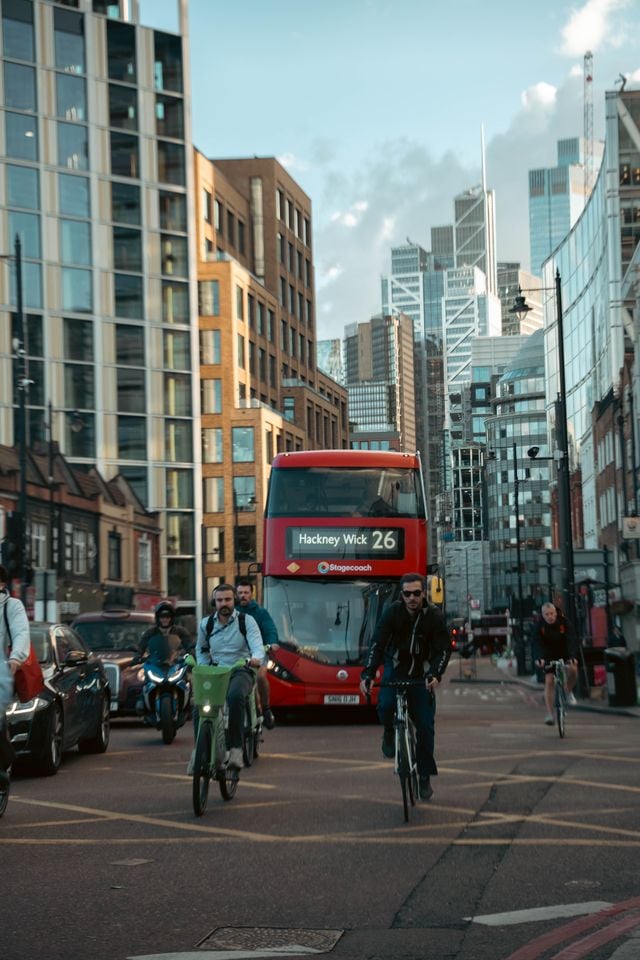 Cyclists ride alongside cars and a red double-decker bus displaying "Hackney Wick 26" on a busy city street, surrounded by tall buildings under a partly cloudy sky—a vibrant scene that captures what living in Hackney is all about.